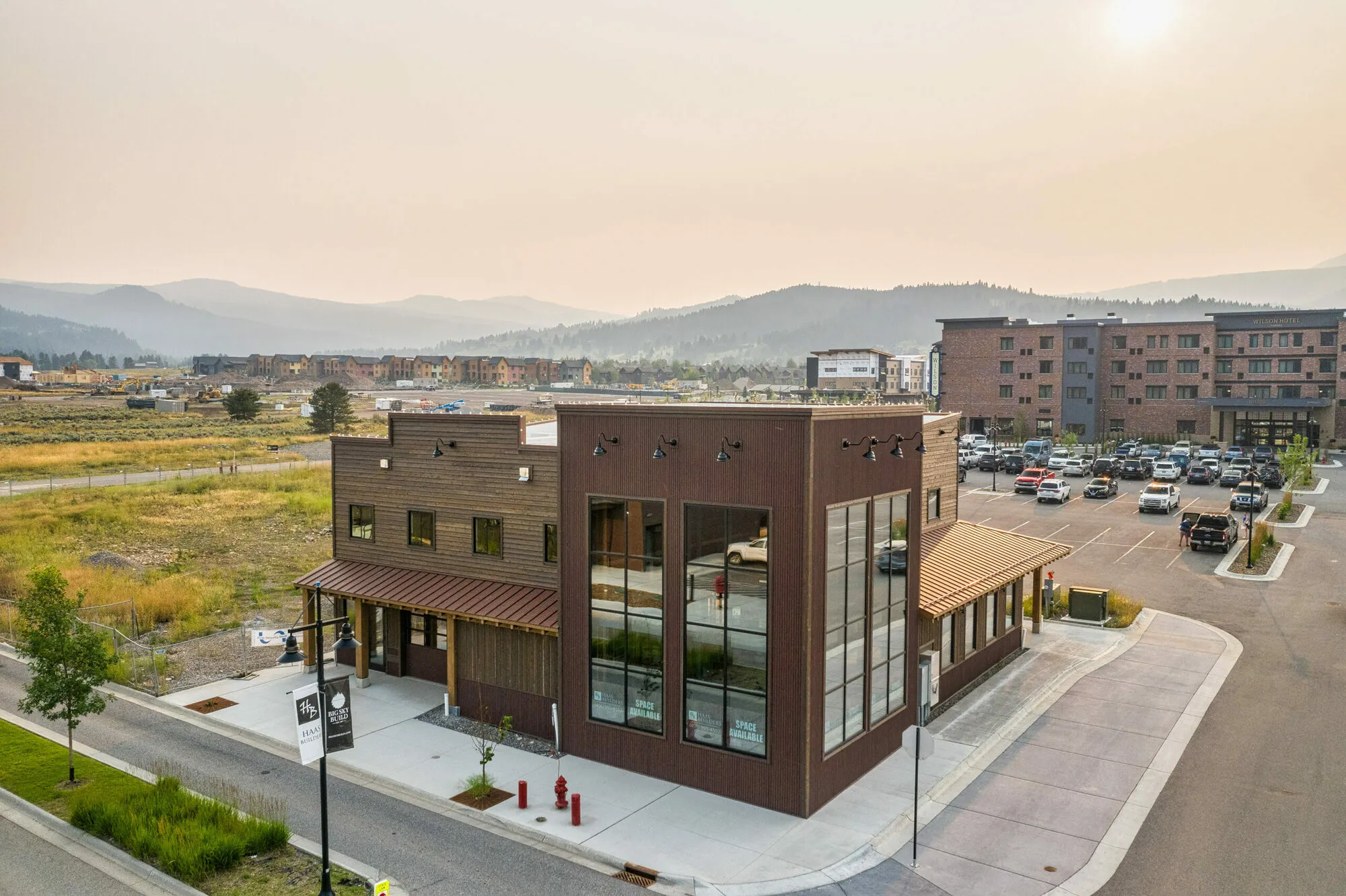 Two-story commercial building with large windows beside a parking lot, with mountains in the background